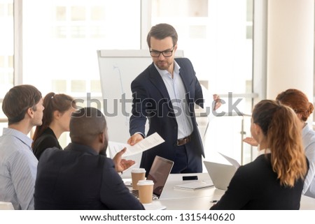 In boardroom gathered multiracial business people in formal wear, confident caucasian coach giving handout or financial report to diverse seminar participants. Information, education teamwork concept Royalty-Free Stock Photo #1315440620