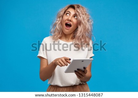 Picture of pretty girl with pinkish hair looking up with amazed fascinated facial expression, opening mouth widely, exclaiming Wow, pointing index finger at screen of digital tablet in her hand