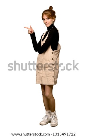 A full-length shot of a Young redhead woman pointing finger to the side in lateral position over isolated white background