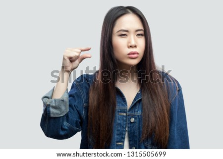 Give me few more. Portrait of beautiful brunette asian young woman in casual blue denim jacket with makeup standing and showing a little size with hand. studio shot, isolated on light grey background.