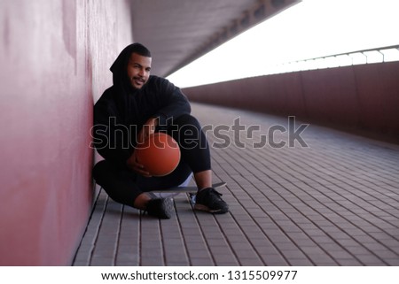 African-American man wearing a black hoodie sitting on a skateboard and holding a basketball while leaning on a wall outside