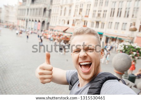 Young bearded blond man smile traveler to Europe with backpack taking selfie on Grand place Belgium Brussels and showing Thumbs up thumb.