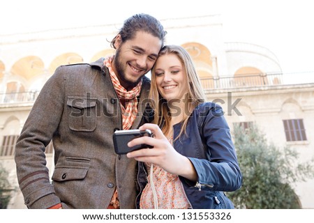 Portrait of a young and fashionable couple taking a picture of themselves while on vacation in a destination city, holding a digital photo camera with their heads together.