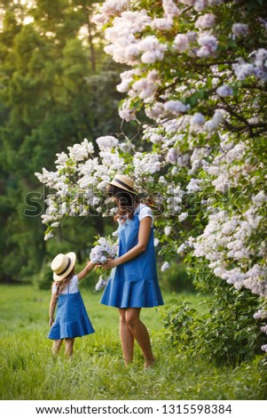 Mother and little daughter walk together in lilac garden. Mom loves her child. Spring story. Family look romantic similar blue dresses and straw hat. Happy family in beautiful spring day