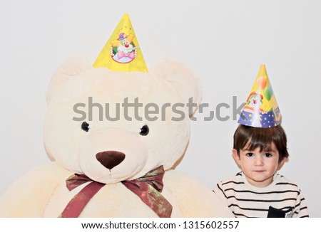 Two years old little boy and his teddy bear best friend wearing birthday hats and posing in front of the camera before kid's birthday party.