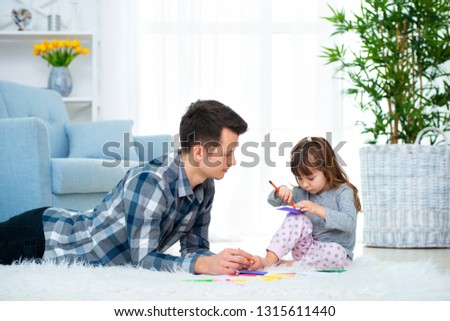 father and little daughter having quality family time together at home. dad with girl lying on warm floor drawing with colorful felt tip pencils
