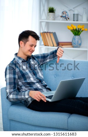 Young guy showing thumb down with his hand at his laptop computer. Man sitting at home on cozy blue sofa at home, wearing casual outfit