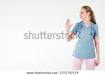 Woman doctor blonde young light gray background studio day beautiful one stethoscope pink purple lilac clothing protests hands forward and keeps feet free stop. Copy space left.