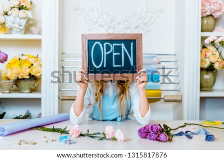 beautiful female flower shop owner holding chalkboard with 'open' lettering in front of face