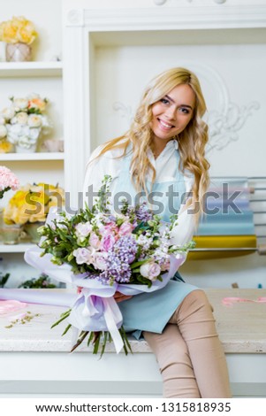 beautiful female flower shop owner looking at camera, sittng on counter and holding bouquet