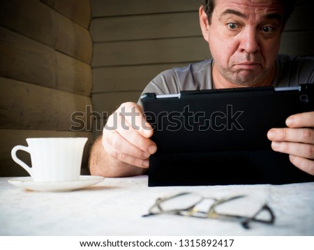 Middle-aged thoughtful man using tablet computer. Book, cup of coffee and glasses on the table