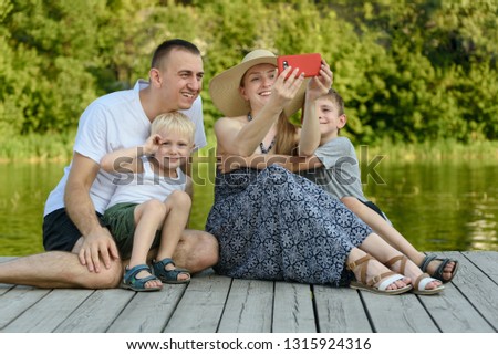 Happy young family, father mother and two little sons are sitting and taking selfies on the river pier.