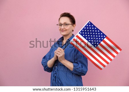 United States flag. Woman holding American flag. Nice portrait of middle aged lady 40 50 years old holding a large USA flag over pink wall background on the street outdoors.