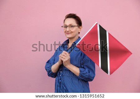 Trinidad and Tobago flag. Woman holding Trinidad and Tobago flag. Nice portrait of middle aged lady 40 50 years old holding a large flag over pink wall background on the street outdoors.