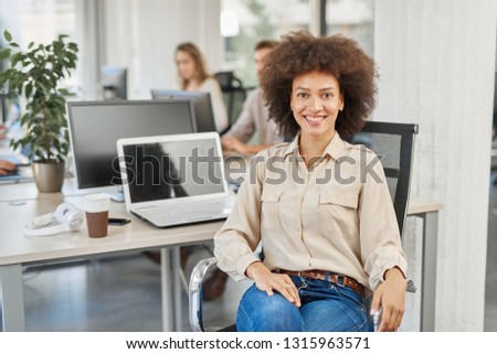 Smiling mixed race CEO sitting at office and looking at camera. In background employees working.
