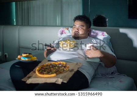 Unhealthy lifestyle concept: Asian obese man eating junk foods while watching TV in bed before sleep Royalty-Free Stock Photo #1315966490