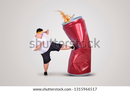 Picture of angry fat man kicking a can of soda drink while wearing sportswear in the studio, isolated on white background