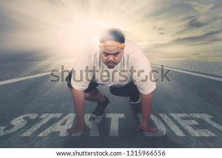 Picture of an young fat man ready to run while kneeling above text of start diet on the road. Shot with sunlight background