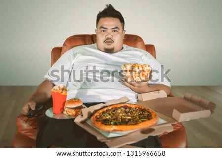 Picture of an obese man watching TV on the sofa while eating junk foods at home 