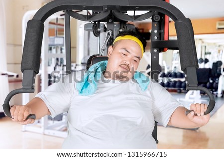 Picture of young obese man looks tired while sitting on the exercise machine. Shot in the gym center