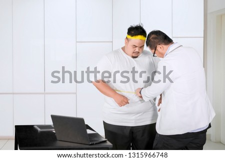 Picture of a male doctor examining fat belly of his patient by using measuring tape. Shot at the clinic