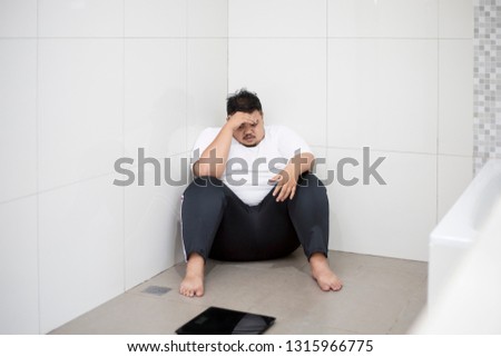 Picture of a frustrated obese man covering his eyes with hands while sitting with scale in the bathroom