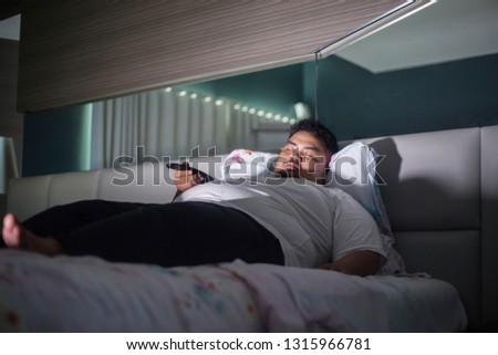 Picture of young fat man holding a remote TV while sleeping on the bed