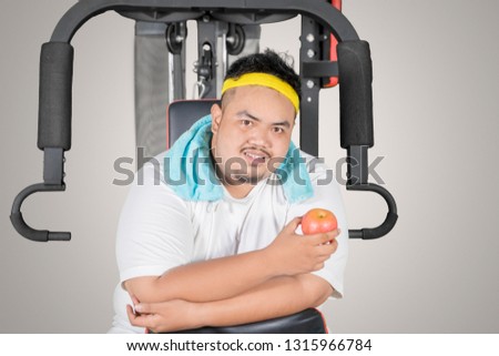 Picture of a young fat man holding a fresh apple while sitting on the exercise machine