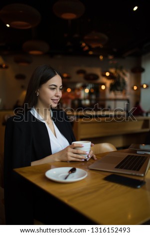 Young gorgeous woman university student having online conference via laptop computer while sitting with cup of coffee in restaurant. Female watching video or movie on notebook, resting in cafe 