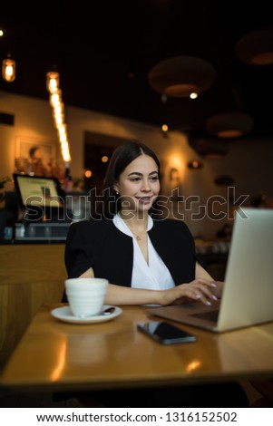 Young beautiful smiling woman successful publication specialist typing article on laptop computer while sitting in coffee shop during recreation time. Happy female copywriter having training course  