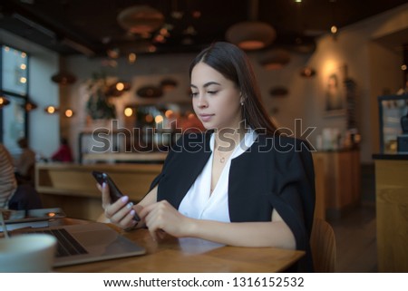 Young fashionable woman blogger reading article on web site via cell telephone while sitting with modern laptop computer in restaurant during recreation time. Female business worker online payment  