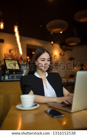 Confident female in formal wear creating summary on website via laptop computer while sitting in restaurant. Woman freelance social media content writer received notifications on netbook 