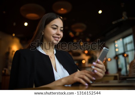 Attractive smiling woman reading pleasant text message on smartphone during leisure time in coffee shop. Happy joyful female manager watching video on cell telephone, sitting in restaurant 