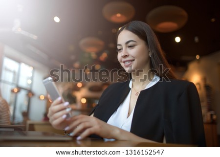 Beautiful smiling business woman sending sms via mobile phone while sitting in modern restaurant during recreation time. Happy joyful female blogger online shopping store via cellphone during resting 