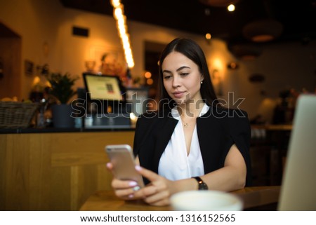 Young successful businesswoman reading e-mail on mobile phone while sitting in restaurant during break at job. Confident female lawyer searching contact on cell telephone 