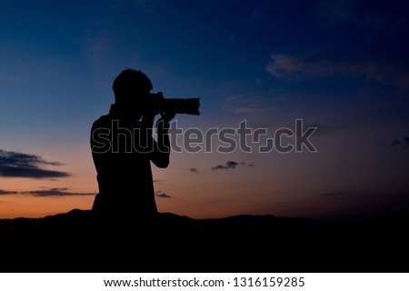 Silhouette of a man who is professional photographer is using a dslr camera at twilight,orange and blue sky on top of the moutain background.Copy space.Ideal for use in documentary, adventure tourism.