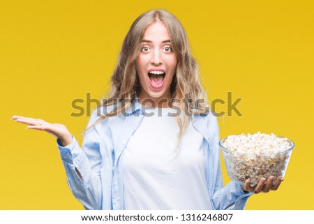 Beautiful young blonde woman eating popcorn over isolated background very happy and excited, winner expression celebrating victory screaming with big smile and raised hands