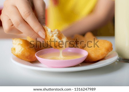 Hand of woman dipping fried buns into sweetened beverage creamer before eat with the glass of soybean milk on the table