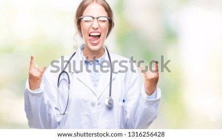 Beautiful young blonde doctor woman wearing medical uniform over isolated background shouting with crazy expression doing rock symbol with hands up. Music star. Heavy concept.