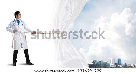 Horizontal shot of young doctor in white medical uniform pulling white fabric while standing over white background with cityscape view behind curtain