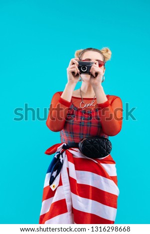 Photo with flag. Stylish blonde-haired woman wearing red bodysuit making photo with American flag