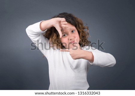 Gesturing finger frame. Portrait of smiling beautiful little girl looking at camera and gesturing finger frame while standing against gray studio background.