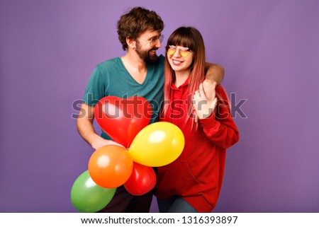 Young loveble hipster couple posing on violet background, bright trendy casual clothes and glasses, hugs and smiling, friendship and relation goals, holding air balloons, ready for celebration.