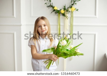 Portrait of a very cute pretty blonde girl in a white shirt against a white wall with a bouquet of white tulips