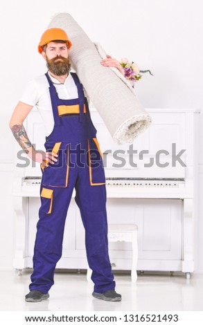 Portrait of man with beard and mustache isolated in white painted room. Builder with tattoo on arm holding rolled carpet on his shoulder.