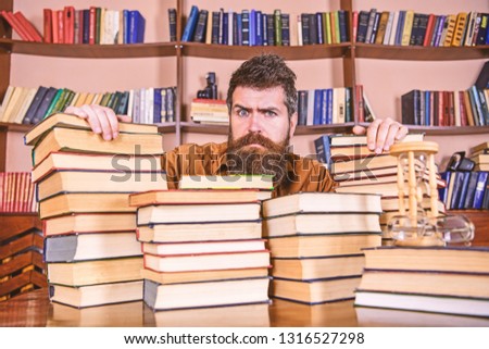 Teacher or student with beard sits at table with books, defocused. Man on serious face between piles of books, while studying in library, bookshelves on background. Bibliophile concept.