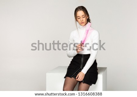 Beautiful girl student in white golf sitting on white cube, holding pink ruler in hand, studio photo on white background