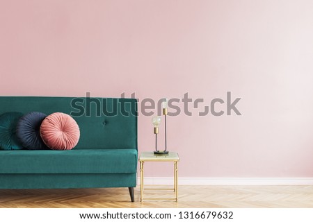 Minimalistic and luxury pastel pink home interior with green velvet design sofa, gold coffe table with table lamp. Copy space for inscription, mock up poster. Empty wall. Brown wooden parquet.