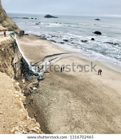 Cloudy Ocean Beach Landscape with Waterfall and People Walking on Sand Nearby