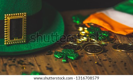 Seasonal display of Saint Patricks Day decor including green leprechaun hat with golden buckle and gold coins, glitter coated shamrocks, and Irish Flag on wooden table top